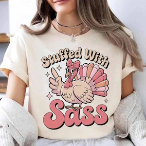 May include: A cream-colored t-shirt featuring a cartoon turkey with a pink bow, pointing a finger. The text "Stuffed With Sass" is written in a retro font. The turkey has a pink wattle and a colorful tail.