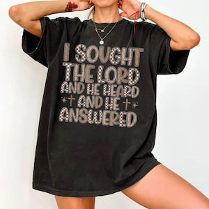 Puede incluir: Camiseta negra de gran tamaño con el texto "I Sought The Lord And He Heard And He Answered" en una mezcla de fuentes marrones y blancas a cuadros y con estampado de leopardo. La camiseta tiene un corte holgado.