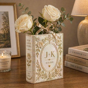 Könnte beinhalten: Eine dekorative, buchförmige Vase mit cremefarbenen Rosen und Grün. Die Vase ist weiß mit goldenen floralen Akzenten und dem Text "OUR LOVE STORY", "J+K" und "10.15.2026". Ein goldenes Band schmückt die Blumen. Eine Kerze und Bücher sind im Hintergrund.