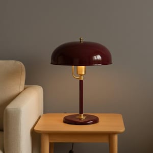 May include: A burgundy-colored desk lamp with a rounded shade and a brass accent. The lamp sits on a small wooden table next to a beige sofa. The lamp's design is a classic, vintage style.