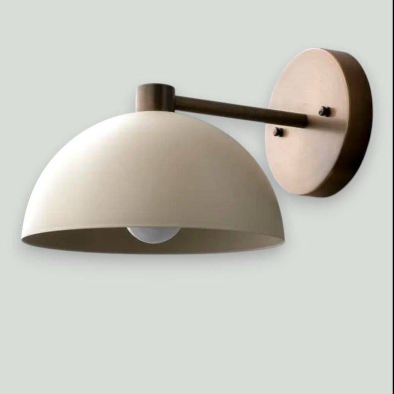 May include: A wall-mounted light fixture with a cream-colored, dome-shaped shade and a bronze-colored arm and mounting plate. The fixture has a simple, modern design, with a visible light bulb.
