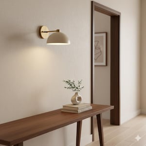 May include: A modern wall sconce with a brass finish and a cream-colored, dome-shaped shade is mounted on a beige wall. A wooden console table holds a small vase with greenery and a stack of books. A doorway is visible in the background.