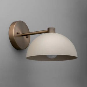 May include: A wall-mounted light fixture with a cream-colored, dome-shaped shade and a bronze-colored metal arm and backplate. The light fixture is designed to be mounted on a wall and provides ambient lighting.