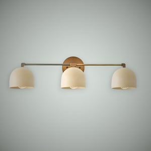 May include: A three-light vanity fixture with a brass finish. The fixture has three cream-colored, dome-shaped shades. The shades are attached to a horizontal bar and a circular backplate.