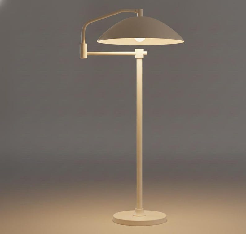 May include: A cream-coloured floor lamp with a curved arm and a dome-shaped shade. The lamp has a circular base and a vertical pole. The light bulb is visible inside the shade. The lamp is made of metal.