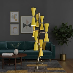 May include: A tall, yellow floor lamp with a brass frame. The lamp has multiple cone-shaped shades and stands on a tripod base. The lamp is in a living room with a teal sofa, a round wooden coffee table, and framed artwork on the wall.