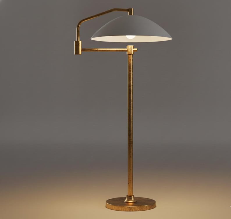 May include: A gold-coloured floor lamp with a white lampshade. The lamp has a curved arm and a round base. The lampshade is a semi-circle shape. The lamp is lit, and the background is a gradient of grey and yellow.
