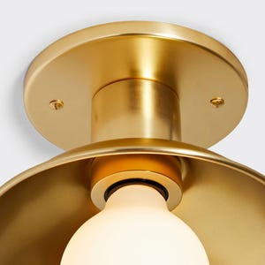 Puede incluir: Primer plano de una lámpara de techo dorada. La lámpara tiene una base redonda y una pantalla curva, con una bombilla visible. La bombilla es blanca y emite un brillo suave. La lámpara es de metal y tiene un acabado pulido.