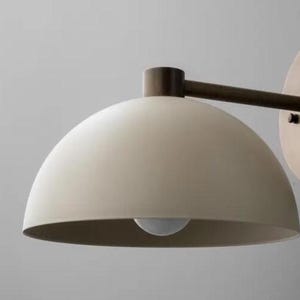 May include: A modern wall-mounted light fixture with a cream-colored, dome-shaped shade. The fixture has a dark bronze arm and a round mounting plate. The light bulb is visible inside the shade. The design is minimalist and contemporary.