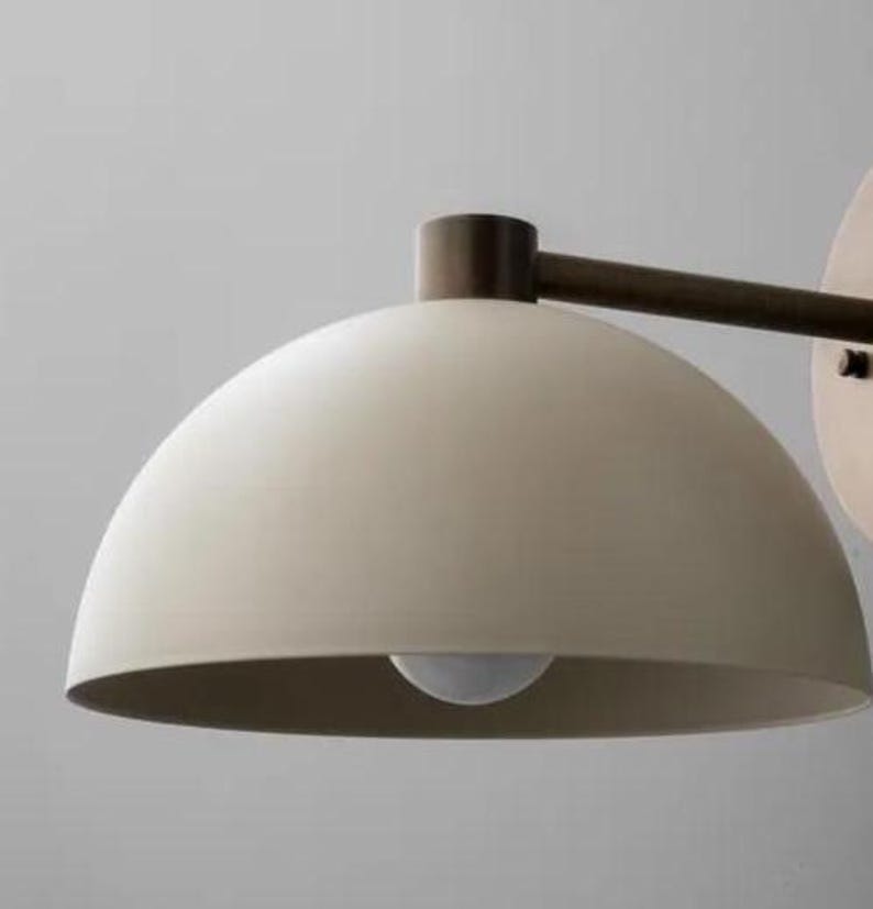 May include: A modern wall-mounted light fixture with a cream-colored, dome-shaped shade. The fixture has a dark bronze arm and a round mounting plate. The light bulb is visible inside the shade. The design is minimalist and contemporary.