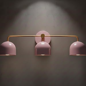May include: A three-light wall sconce with a pink finish. The light fixture has a gold-colored horizontal bar and a circular wall mount. Each light has a rounded, dome-shaped shade. The sconce is mounted on a dark gray wall.