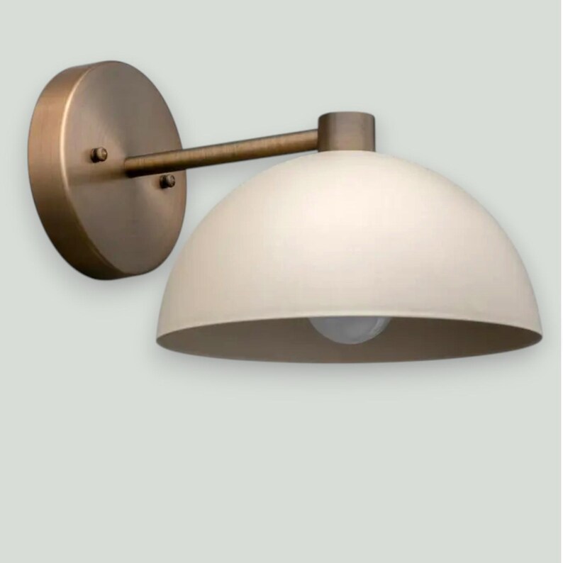 May include: A wall-mounted light fixture with a circular, bronze-colored base and a curved arm supporting a cream-colored, dome-shaped shade. The fixture includes a visible light bulb. Ideal for home decor and interior design.