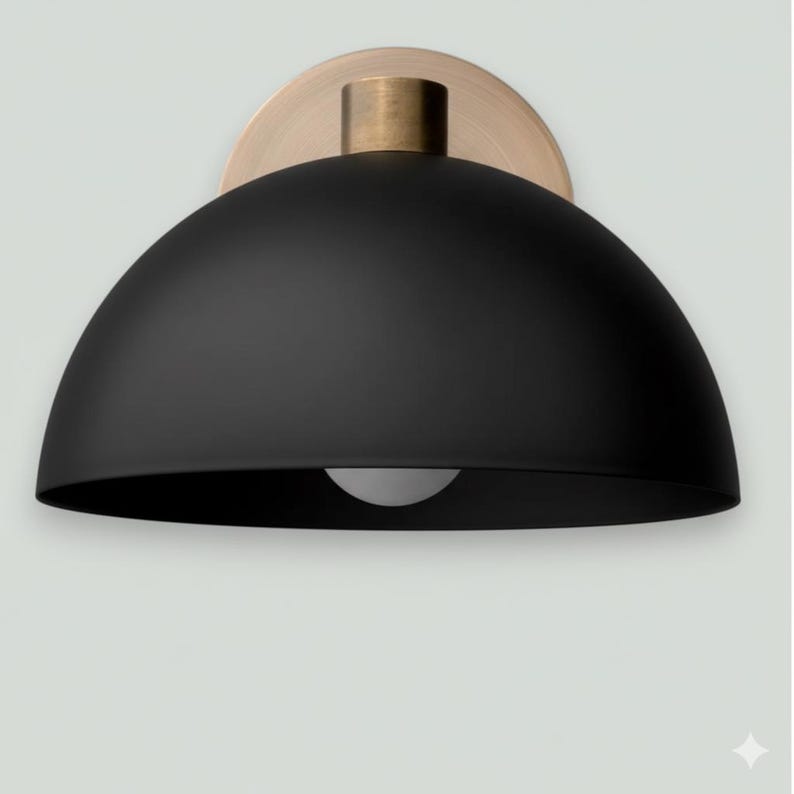 May include: A black dome-shaped wall sconce with a brass-colored mounting plate and a cylindrical accent. The sconce features a single light bulb. The design is modern and minimalist.