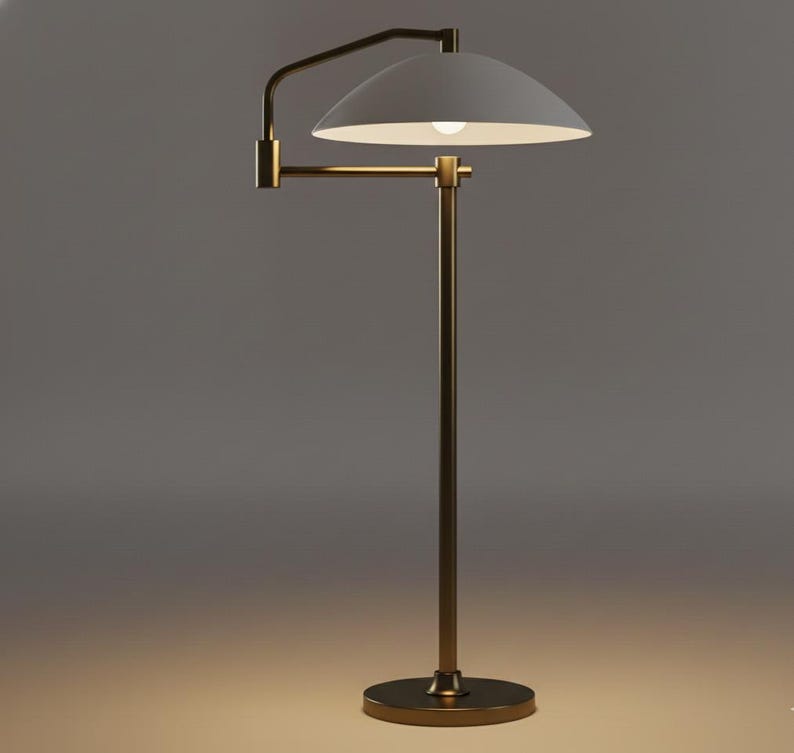 May include: A contemporary floor lamp featuring a white, semi-circular lampshade and a brass-toned, adjustable arm. The lamp has a tall, slender brass pole and a circular base. The light bulb is visible inside the shade. Designed for indoor use.