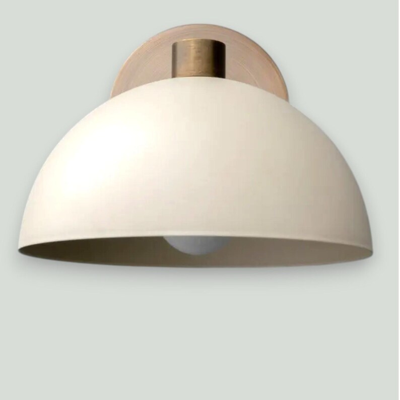 May include: Cream-colored dome-shaped wall sconce with a brass-colored mounting plate and a cylindrical fixture. The light fixture has a simple, modern design, suitable for various interior styles. The light bulb is visible at the bottom of the dome.