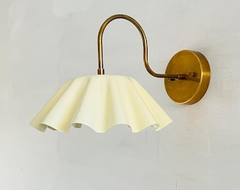 May include: A wall-mounted light fixture with a cream-colored, ruffled lampshade and a curved brass arm. The fixture has a round brass base and a simple, elegant design, suitable for interior lighting.