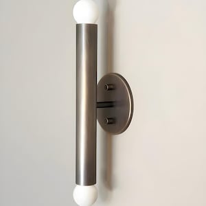 May include: A modern wall sconce with a cylindrical, brushed metal body and a circular mounting plate. The sconce features two white globe-shaped light bulbs, one at the top and one at the bottom of the cylinder. The fixture is designed for indoor use.