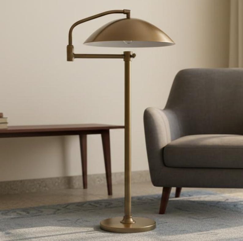 May include: A brass floor lamp featuring a domed shade and an adjustable arm. The lamp has a circular base and a straight pole. The shade is positioned above a grey armchair. The lamp is in a room with a wooden table and a patterned rug.