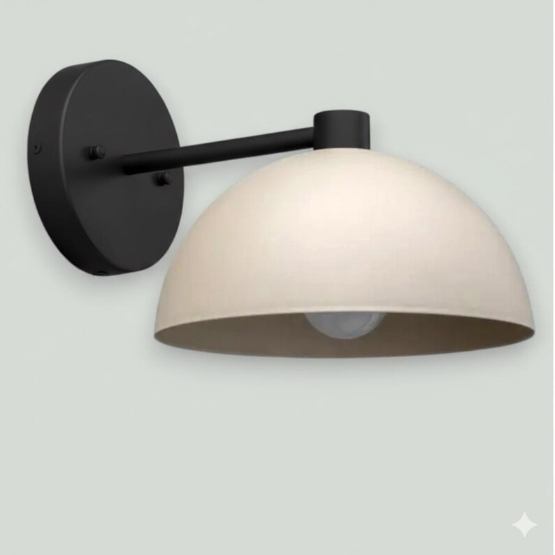 May include: A modern wall sconce with a matte black finish and a cream-colored, dome-shaped shade. The light fixture has a circular base and a straight arm extending to the shade, which houses a light bulb.