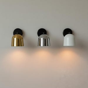 May include: Three contemporary wall lights, each with a black circular mount and a bell-shaped shade. The shades are gold, silver, and white. The lights are fixed to a light-coloured wall, with a warm glow beneath each.