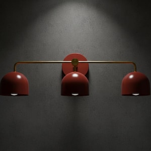 May include: A modern wall-mounted light fixture with three dome-shaped shades in a deep red color. The shades are suspended from a gold-colored horizontal bar, attached to a circular red base. The fixture is designed for interior lighting.
