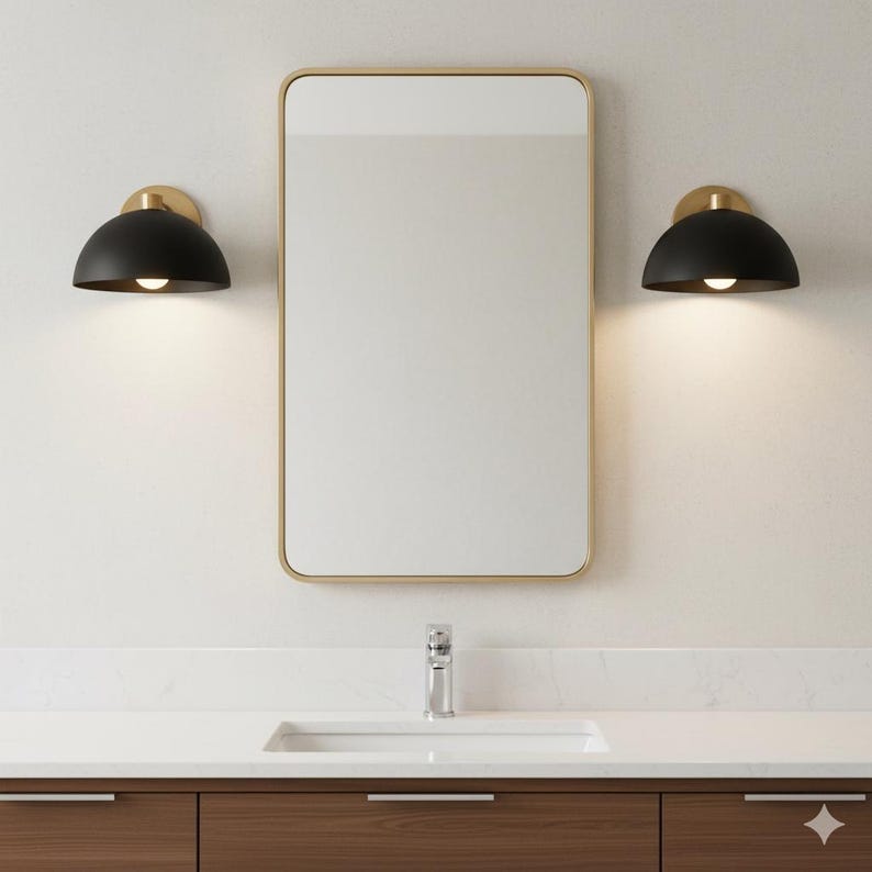 May include: Bathroom vanity with a rectangular mirror in a gold frame, two black dome-shaped sconces with gold accents, and a white countertop with a sink and faucet. The vanity has dark wood cabinets.
