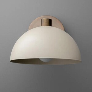 May include: Cream-colored, dome-shaped wall sconce with a brass-colored stem and mounting plate. The light fixture has a simple, modern design, and the bulb is visible from below. The sconce is mounted on a gray wall.