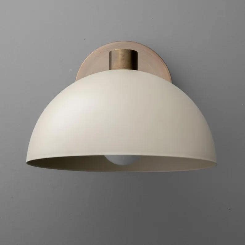 May include: Cream-colored, dome-shaped wall sconce with a brass-colored stem and mounting plate. The light fixture has a simple, modern design, and the bulb is visible from below. The sconce is mounted on a gray wall.