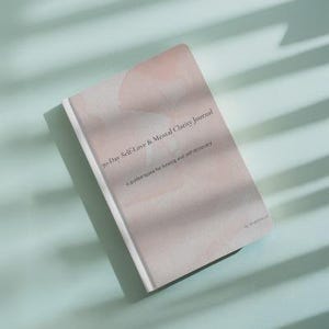 May include: A journal titled "30-Day Self-Love & Mental Clarity Journal" with a soft pink and white cover. The journal is lying on a light blue surface with shadows. The text on the cover reads "A guided space for healing and self-discovery."