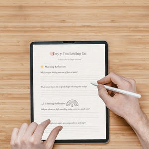 May include: A digital tablet displaying a journal page titled "Day 7: I'm Letting Go." The page includes prompts for morning and evening reflections. A hand is using a white stylus to write on the screen. The tablet rests on a wooden surface.