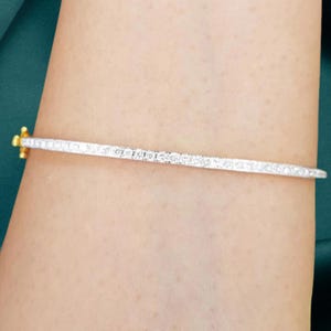 Könnte beinhalten: Ein silberfarbenes Armband mit einem Schwertdesign. Das Armband hat eine dünne, längliche Form mit strukturierter Oberfläche. Der Knauf des Schwertes ist goldfarben. Das Armband wird am Arm getragen.