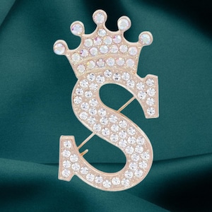 Peut inclure: Broche en métal doré en forme de lettre "S" surmontée d'une couronne. La broche est incrustée de nombreux cristaux clairs et étincelants. L'épingle est posée sur un fond vert foncé.