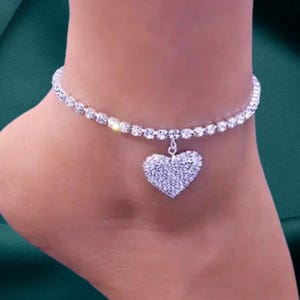 May include: A silver anklet with a heart-shaped pendant. The anklet is adorned with a row of small, clear crystals. The heart pendant is also covered in clear crystals. The jewellery is worn on an ankle.
