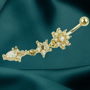 May include: A gold-colored belly button ring with a dangling floral design. The ring features a small ball at the top and a series of clear, sparkling gemstones arranged in a flower and teardrop shape. The jewelry is set against a dark teal background.