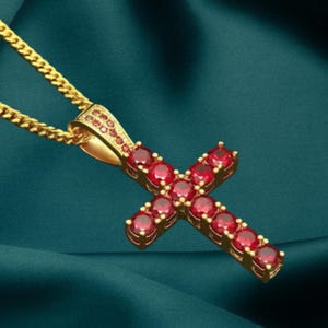 Puede incluir: Un collar con colgante de cruz dorada y cadena. La cruz está adornada con numerosas piedras preciosas rojas. El fondo es de tela verde oscuro.