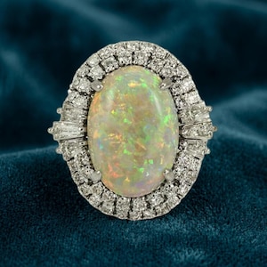 May include: An oval-shaped cabochon opal ring featuring a prominent play-of-color with flashes of green, orange, and yellow. The center stone is surrounded by a double halo of small round brilliant-cut diamonds and baguette-cut diamond accents.