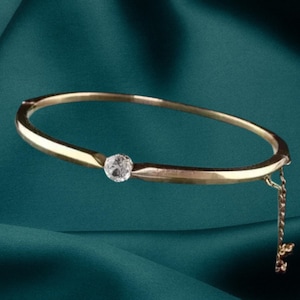 Puede incluir: Un brazalete de oro con una sola gema clara y redonda en el centro. Una delicada cadena de oro con un pequeño dije cuelga del brazalete. El brazalete está sobre un fondo verde azulado.