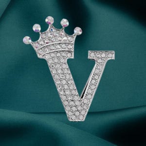 May include: A silver-toned brooch in the shape of the letter "V" topped with a crown. The brooch is encrusted with clear, sparkling rhinestones. The crown has iridescent, pearlescent stones at the tips.