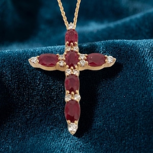 Puede incluir: Un colgante de cruz de oro con cinco rubíes de color rojo intenso en talla oval dispuestos en un patrón vertical y horizontal, acentuado por pequeños diamantes blancos redondos engastados entre los rubíes, suspendido de una fina cadena de oro.