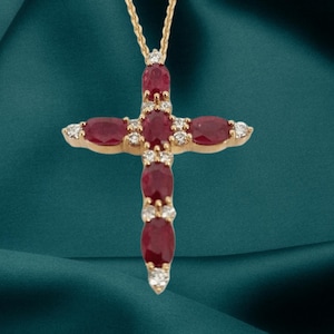 Pode incluir: Colar com pingente de ouro com design de cruz. A cruz é adornada com pedras preciosas vermelhas de corte oval e pequenas pedras preciosas redondas e transparentes. O colar pende de uma fina corrente de ouro.