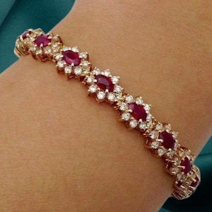 Può includere: Bracciale in oro con pietre preziose rosse taglio ovale e piccole pietre preziose chiare e rotonde. Il design del bracciale include gruppi a forma di fiore, con le pietre rosse al centro. Il bracciale è su un polso.