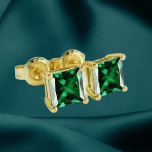 May include: A pair of gold-coloured stud earrings featuring square-cut, emerald-green gemstones. The earrings are set in gold-coloured metal and have a post and backing. The gemstones are a vibrant green colour.