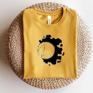 May include: Mustard yellow t-shirt with a black and white graphic design. The design features a stylized face within a black blotch. The shirt is folded and placed on a woven surface. The text "AI SVG EPS DXF PNG" is on the left.