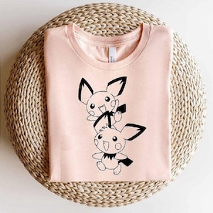 May include: A light pink t-shirt featuring a black and white cartoon graphic of two characters with large ears and smiling faces. The shirt is folded neatly on a woven, circular surface. The image also includes the text "AI SVG EPS DXF PNG".