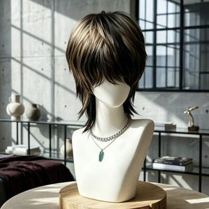 Puede incluir: Una peluca de capas a la altura de los hombros con reflejos castaños oscuros y rubios en un corte desfilado. La peluca se exhibe en una cabeza de maniquí blanca que lleva un collar de cadena plateada con un pequeño colgante verde en forma de hoja.