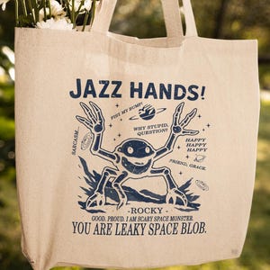 May include: A beige canvas tote bag with the words "JAZZ HANDS!" and a blue cartoon space monster graphic. The bag is filled with white flowers. Text on the bag includes "Sarcasm...", "Why stupid, question?", and "You are leaky space blob."