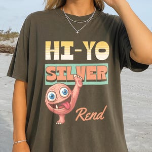 Könnte beinhalten: Ein dunkelgraues Kurzarm-T-Shirt mit einem Grafikdruck, der den Text HI-YO SILVER und Rend zeigt. Das Design umfasst einen gelb-weiß gestreiften Balken im Retro-Stil, einen türkisfarbenen rechteckigen Block und ein rosa Cartoon-Wesen mit großen blauen Augen.