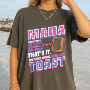Könnte beinhalten: Ein dunkelgraues T-Shirt mit einem Grafikdruck, der den Text "MANA TOAST. THIS IS TOAST. IT REFILLS YOUR MANA. THAT'S IT. NOTHING MORE. FUCK YOU. TOAST" in verpixelter rosa und blauer Schrift sowie eine Illustration von Toastbrot zeigt.