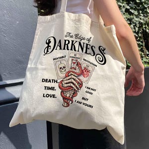 May include: Cream-colored tote bag with the text "The Edge of Darkness" and illustrations of tarot cards, a skull, and a snake wrapped around a skeletal hand. The bag also features the phrases "Death, Time, Love" and "I am not good but I am yours."
