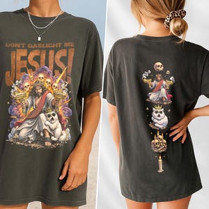 K&ouml;nnte beinhalten: Dunkelgraues T-Shirt mit einer Grafik von Jesus, der Waffen h&auml;lt, umgeben von Totenk&ouml;pfen und dem Text "DON'T GASLIGHT ME JESUS!". Die R&uuml;ckseite des Shirts zeigt ein vertikales Design mit Jesus, einer Katze und Totenk&ouml;pfen.